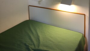 Una habitación pequeña con cama individual, colcha verde, cabecera de madera y dos lámparas fijadas a la pared.