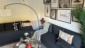 Un salon moderne avec un canapé gris foncé, une table basse ronde ornée d’un vase de fleurs et un mur décoré de tableaux encadrés, ainsi qu’un luminaire au plafond au design original.