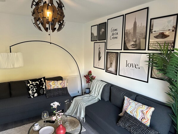 Un salon moderne avec un canapé gris foncé, une table basse ronde ornée d’un vase de fleurs et un mur décoré de tableaux encadrés, ainsi qu’un luminaire au plafond au design original.