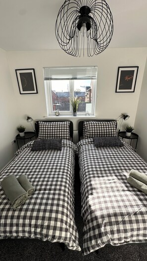 Een slaapkamer met twee bedden, een raam met zonwering, een hanglamp en ingelijste foto's aan de muur.