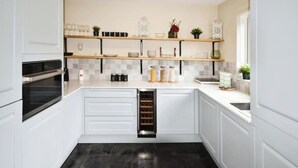 Une cuisine moderne avec des meubles blancs, un four encastré, un réfrigérateur à vin et des étagères décoratives où sont exposés de la verrerie et des plantes.