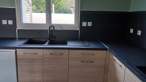 A kitchen with a black countertop, a double sink, wooden cabinets, and a window with a view of greenery outside. - Charming new house with terrace and garden (Muron)