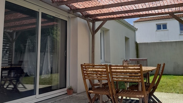 A covered patio with a wooden table and chairs, a sliding glass door, and a pergola with a patterned roof. - Charming new house with terrace and garden (Muron)
