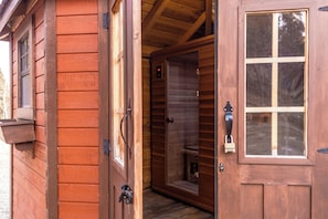 Uma cabana de madeira com porta e janela também de madeira, um pequeno alpendre e uma porta interior visível.