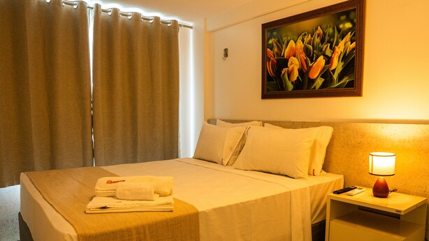 A hotel room with a bed, a nightstand with a lamp, and a painting on the wall. - Magnific (João Pessoa)