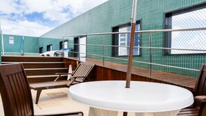 A covered outdoor seating area with wooden chairs and a white table, overlooking a building with green-tiled walls and windows. - Magnific (João Pessoa)