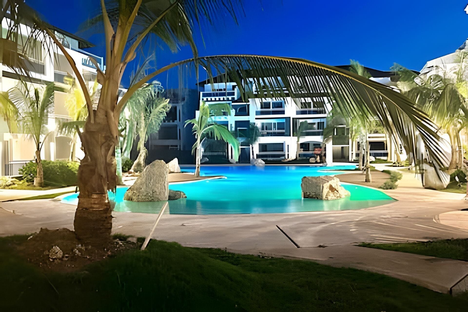 Ein modernes Hotel mit großem Schwimmbad, Palmen und einer überdachten Liegefläche.