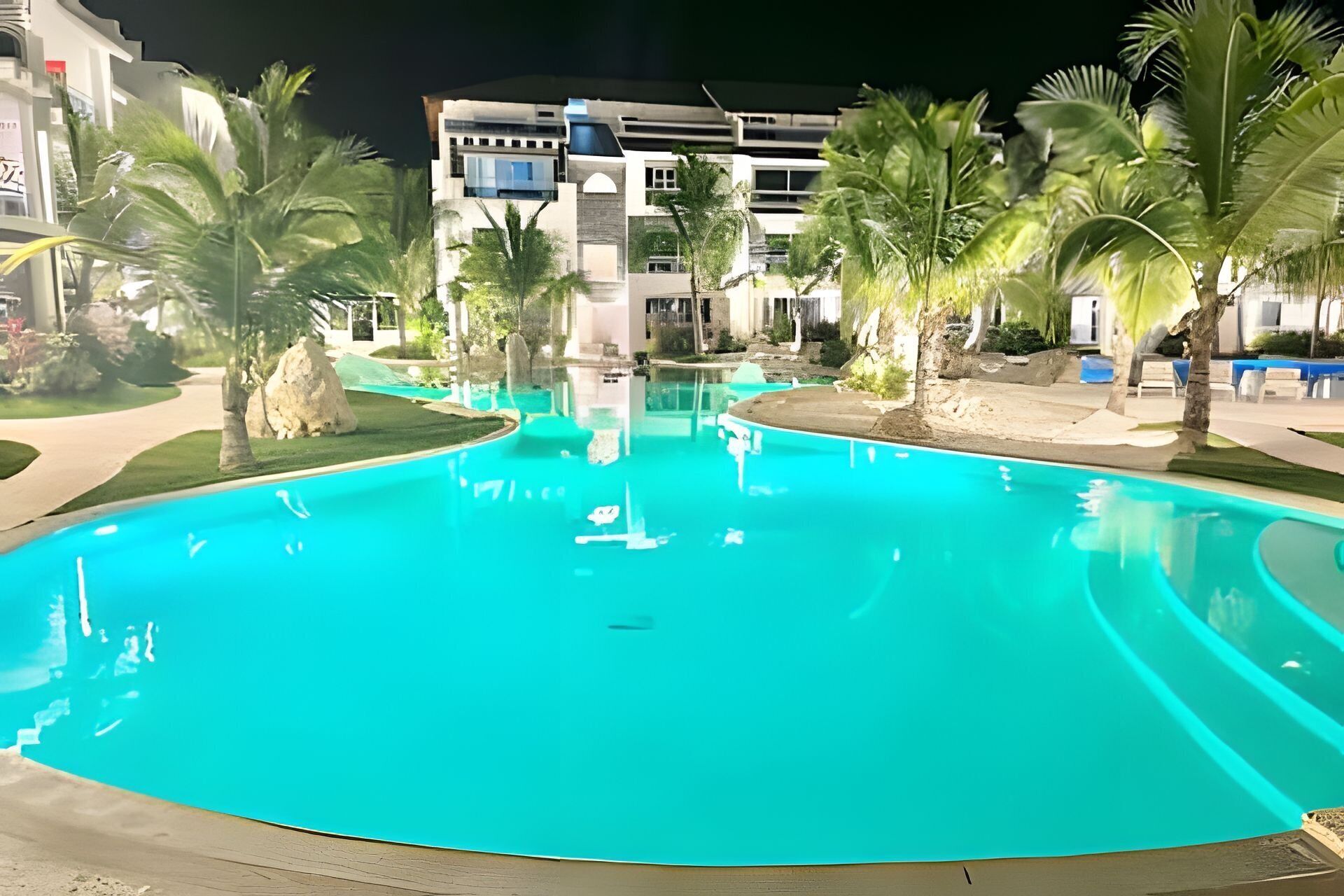 A swimming pool with a curved edge, surrounded by palm trees and a building in the background.