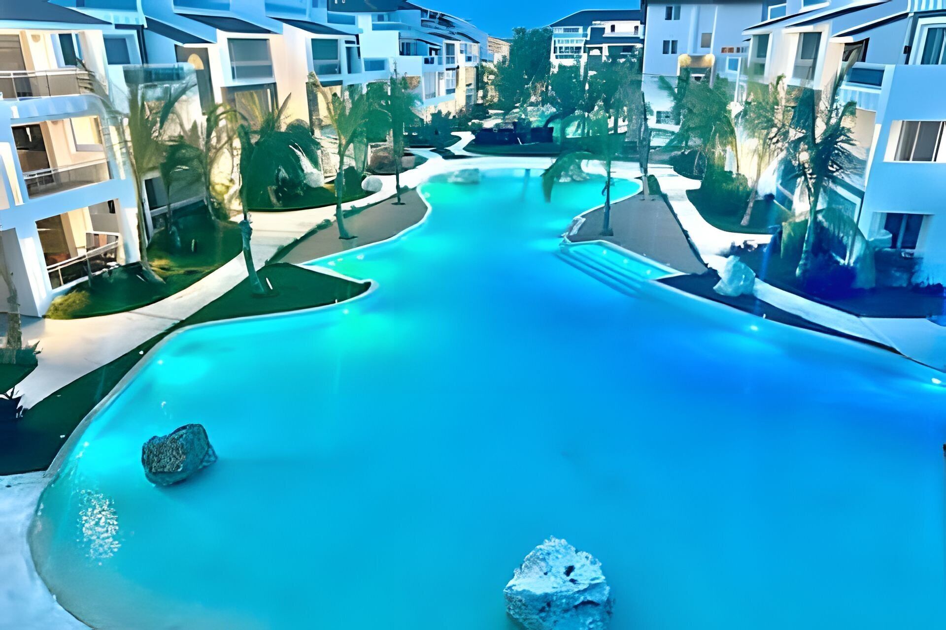 A resort with a central swimming pool, surrounded by modern buildings and palm trees.