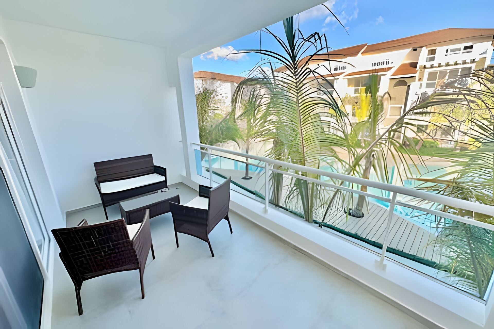 A balcony with a seating area, a glass railing, and a view of a building with palm trees and a pool.