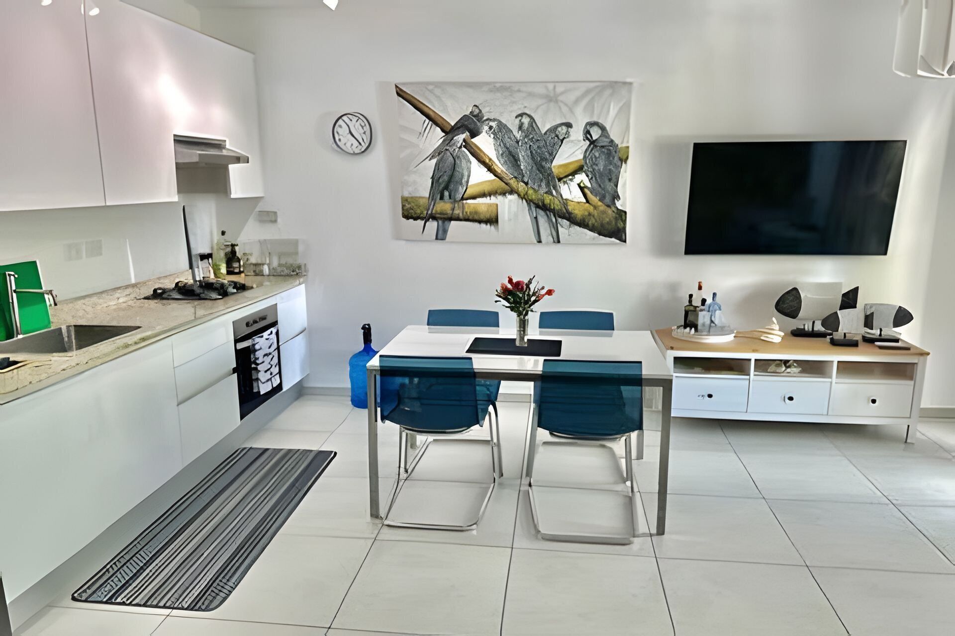 Una cucina moderna con mobili bianchi, un'area pranzo con sedie blu e un grande quadro di uccelli appeso al muro.