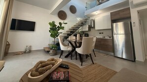 A modern kitchen with a stainless steel refrigerator, a dining area with a glass table and chairs, and a staircase leading to an upper level.