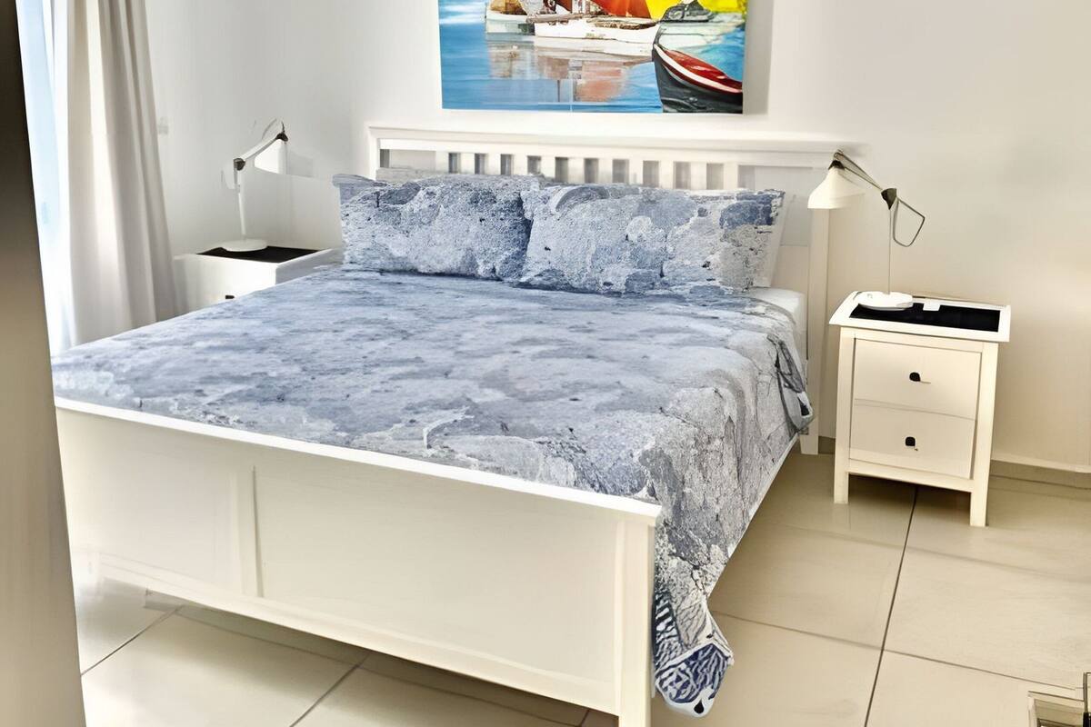 A bedroom with a bed, bedside tables, a painting of boats on water, and a window with curtains.