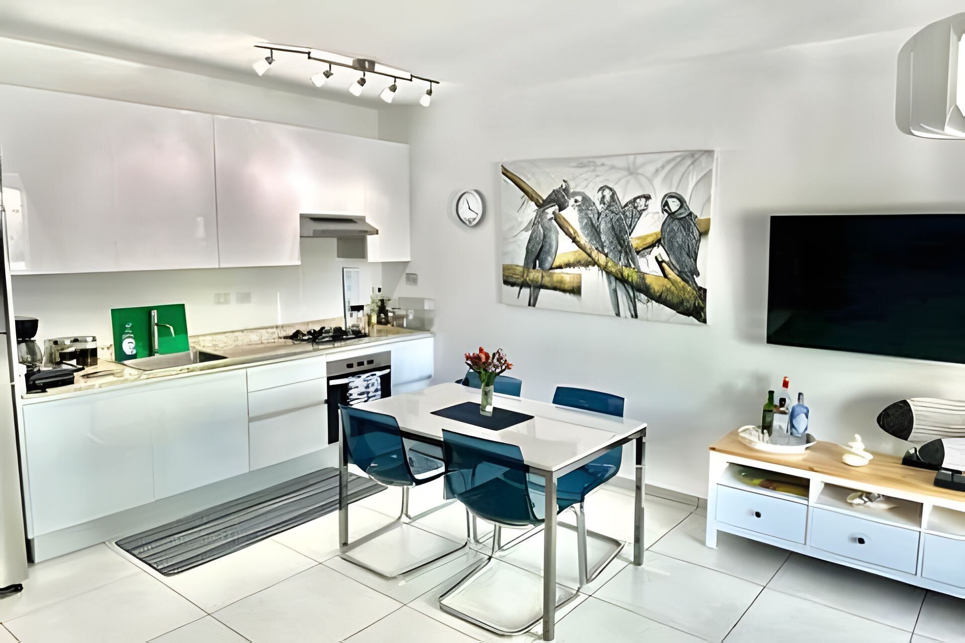 A modern kitchen with white cabinets, a dining area with blue chairs, and a flat-screen TV on the wall.