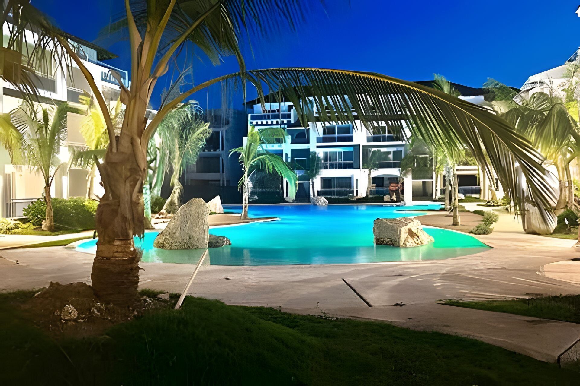 A swimming pool with a modern building in the background, surrounded by palm trees and rocks.