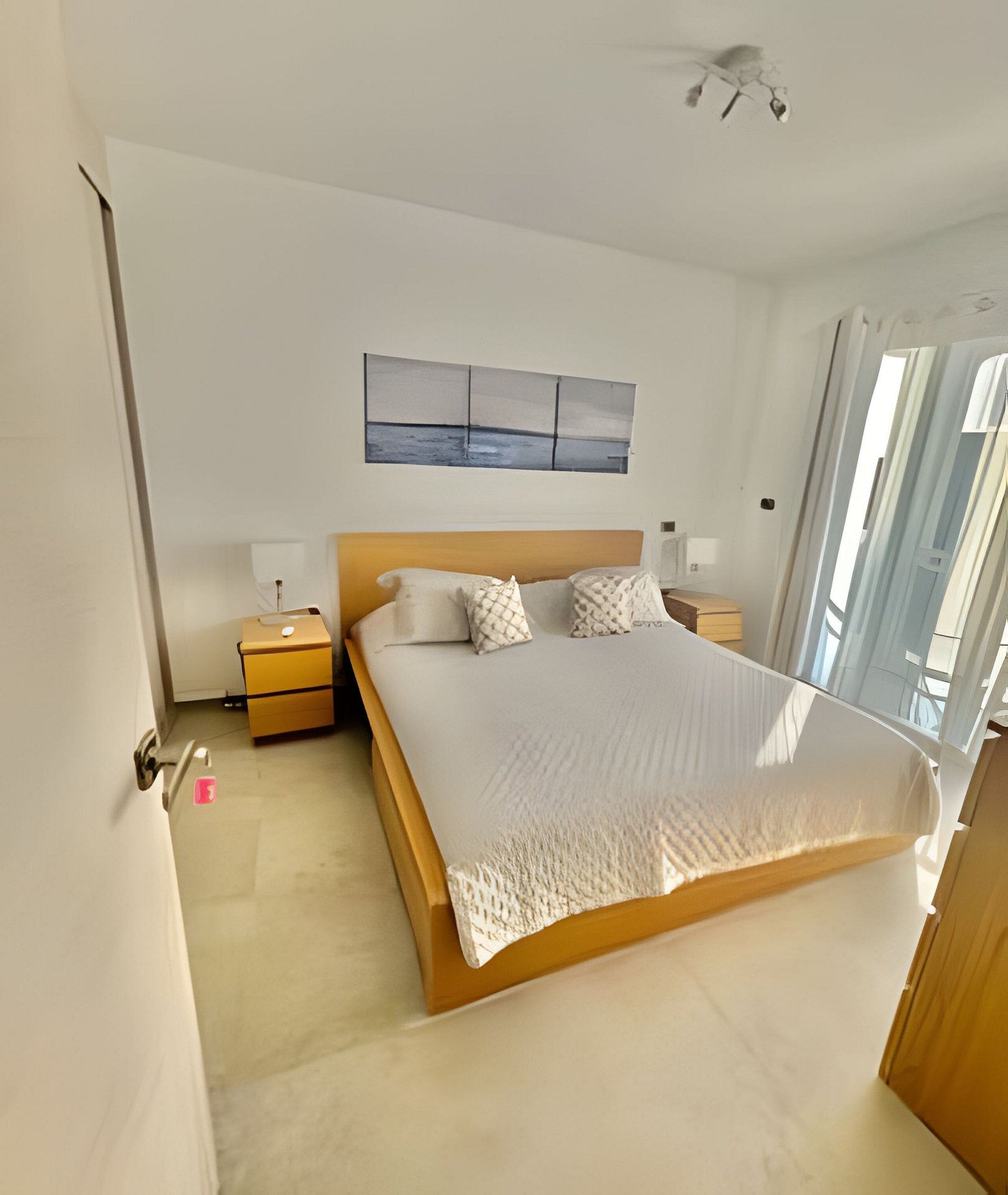 Un dormitorio con cama, mesita de noche, lámpara de pared y ventana con cortinas.