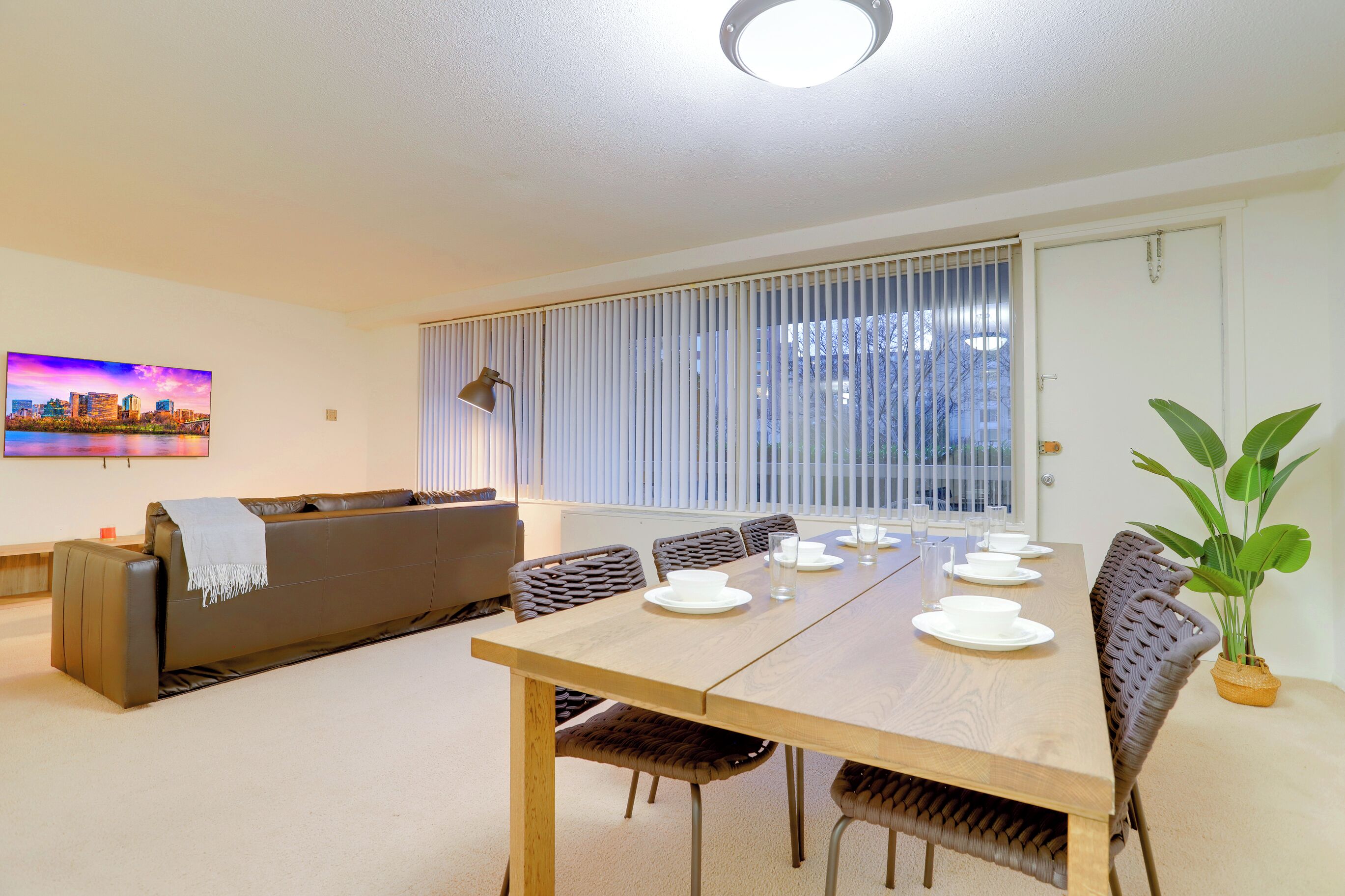 Un coin repas avec une table en bois dressée pour quatre personnes, entourée de chaises en osier, d’un canapé et d’une plante en pot.
