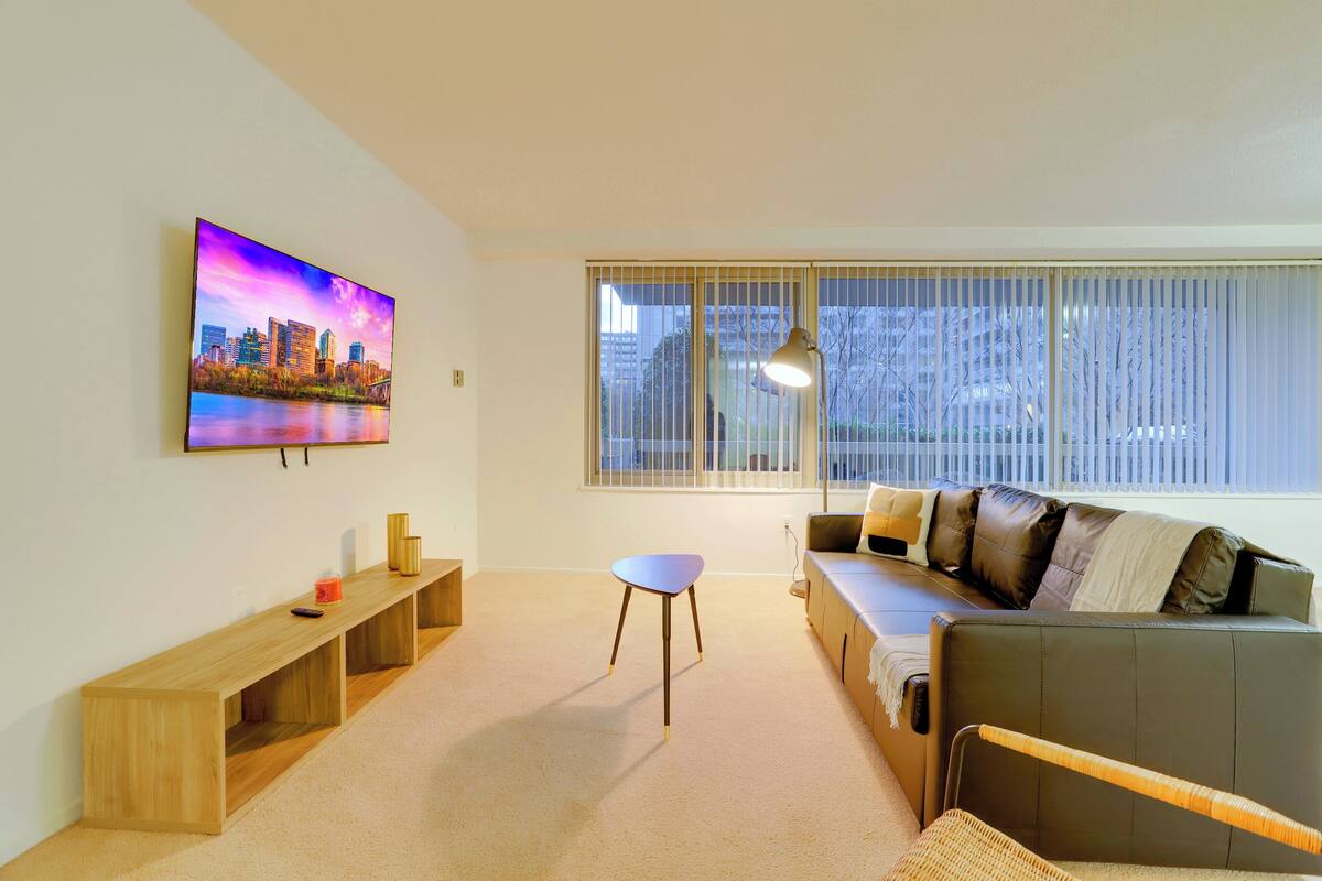 A modern living room with a large flat-screen TV, a sofa, a small table, and a window with blinds.