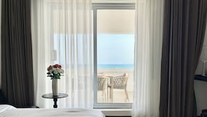 A bedroom with a bed, a small table with a vase of flowers, and a view of the beach through a glass door. - Joy Beach Lodge (Sekondi-Takoradi)
