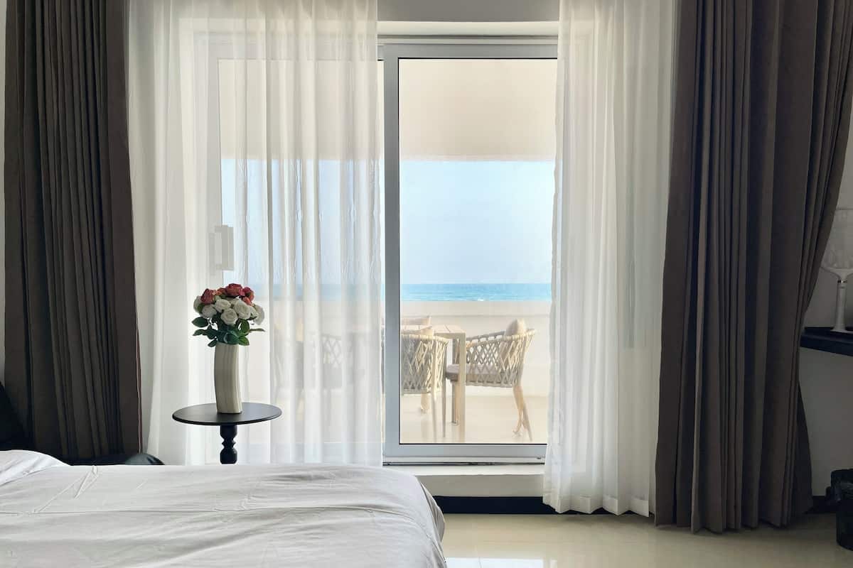 A bedroom with a bed, a small table with a vase of flowers, and a view of the beach through a glass door.