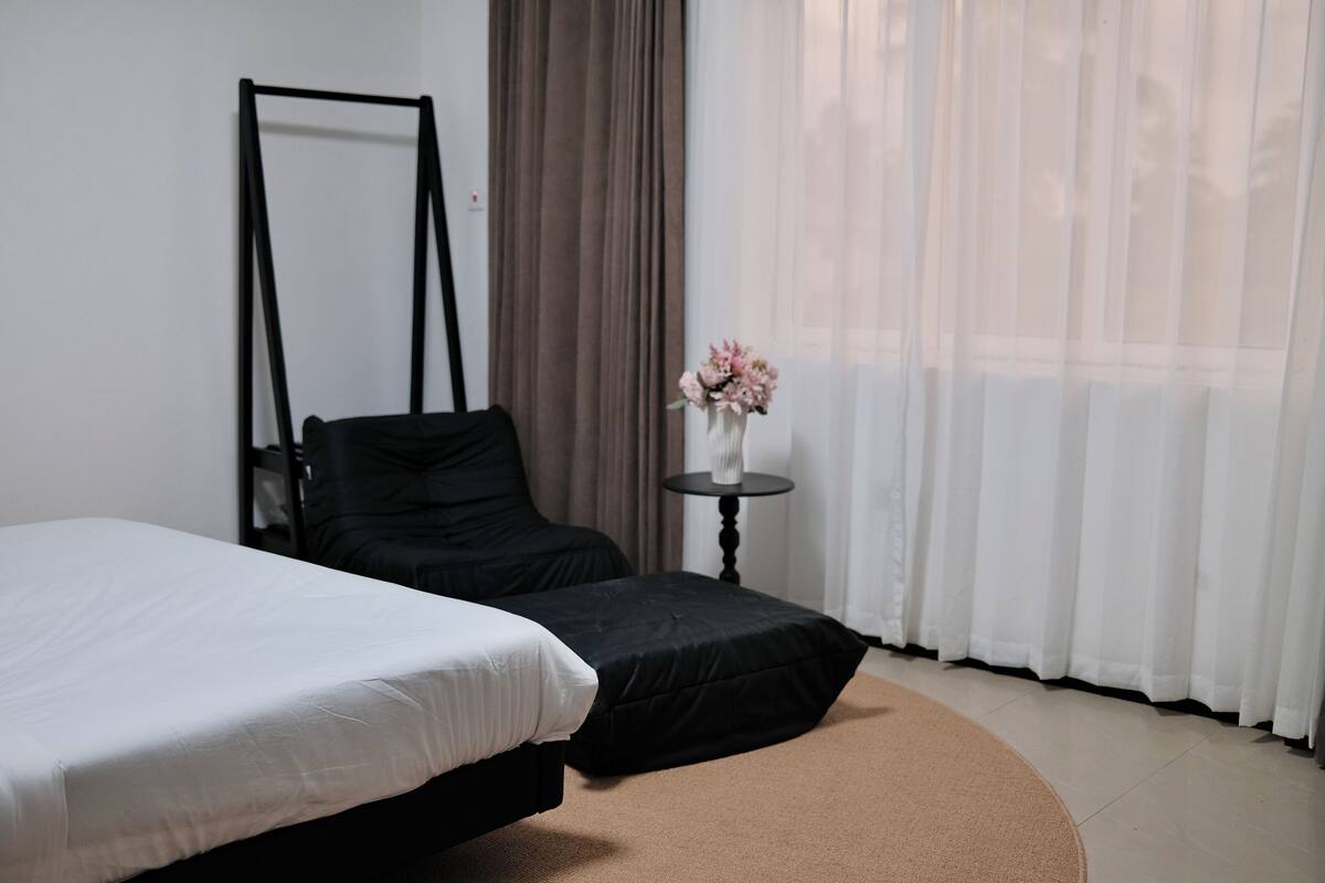 A bedroom with a bed, a chair, a small table with a vase, and a window with curtains.