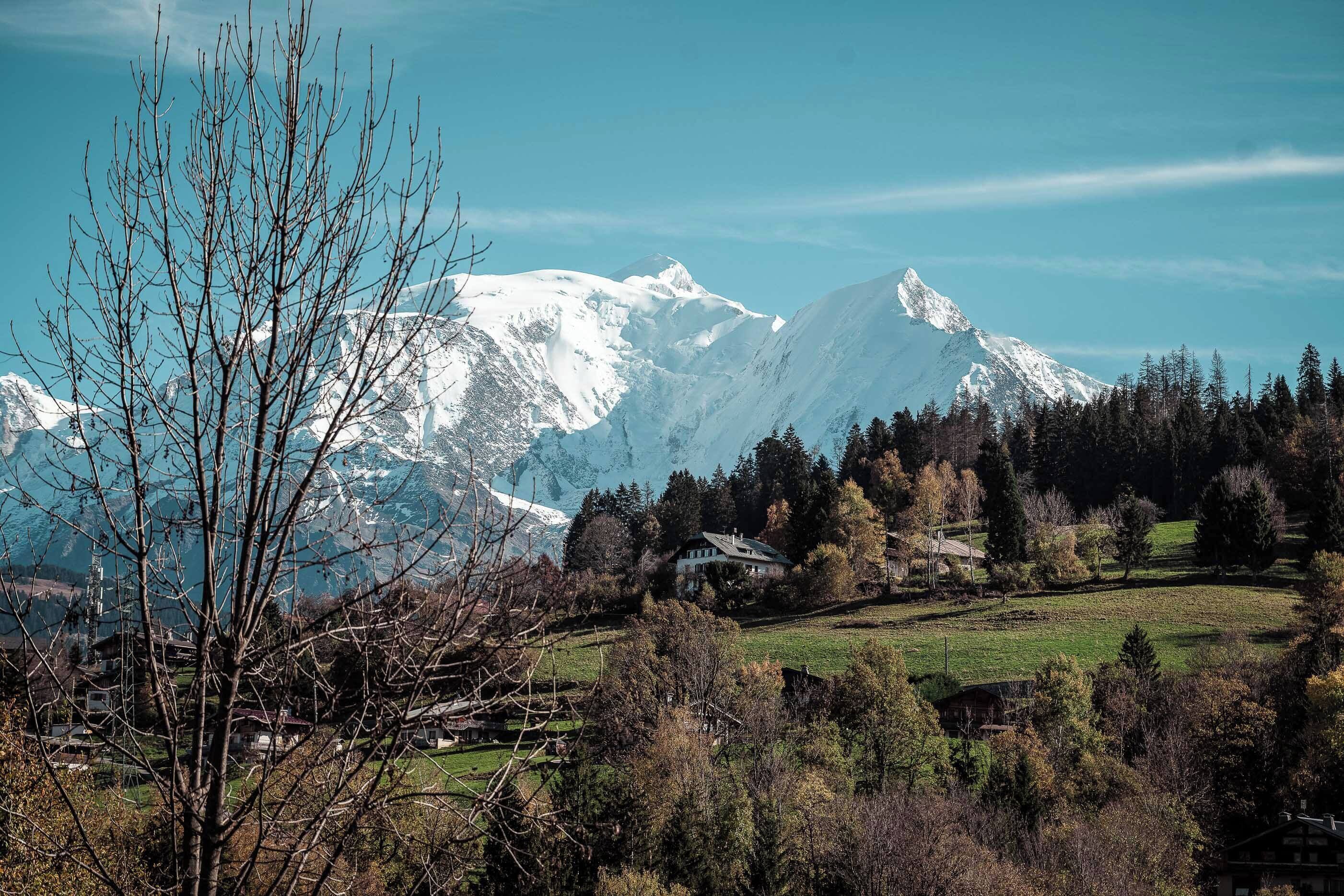 Een bergachtig landschap met besneeuwde toppen, een heldere lucht en op de voorgrond bomen en gebouwen.
