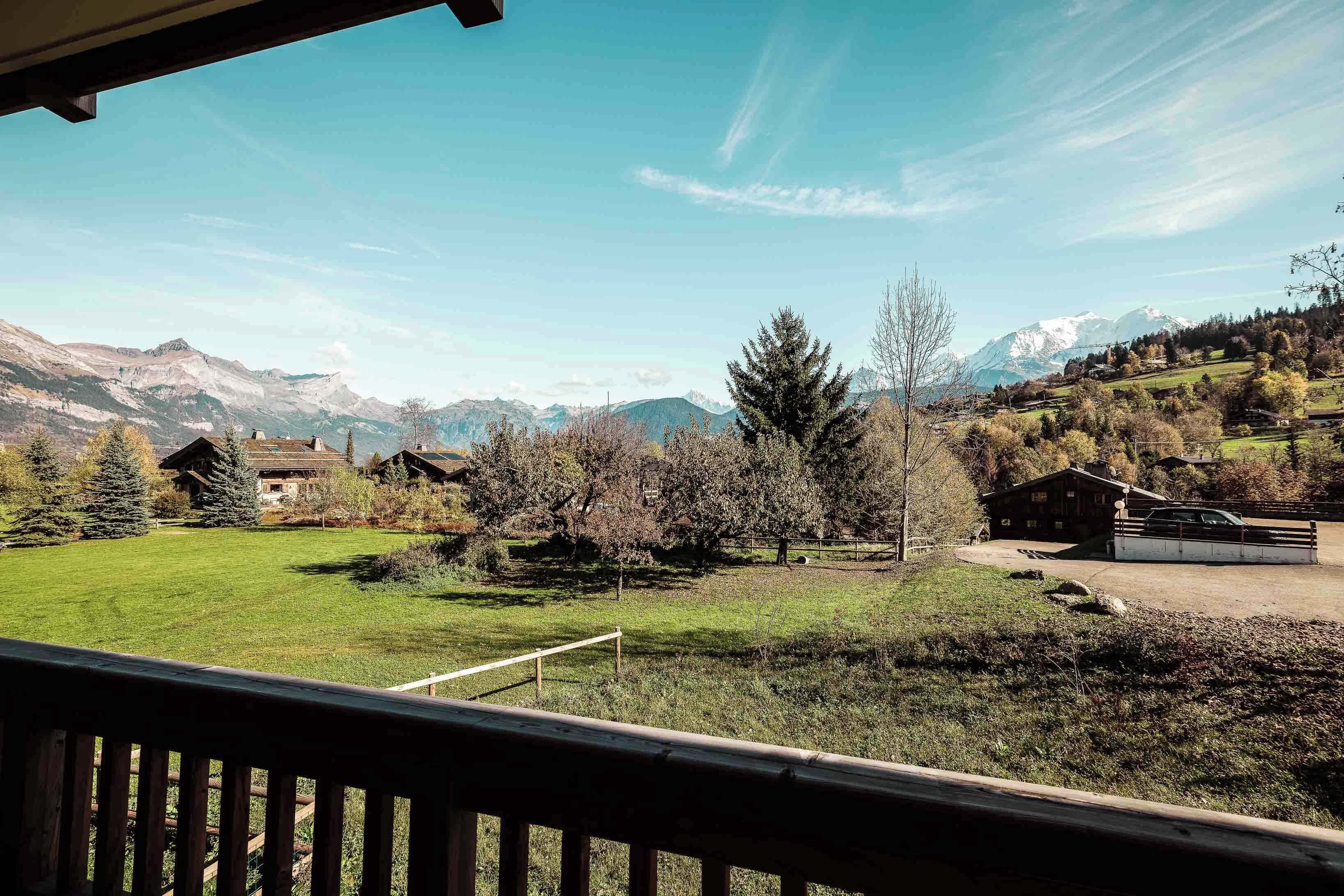 Blick vom Balkon auf ein grünes Feld mit Häusern und Bergen im Hintergrund.