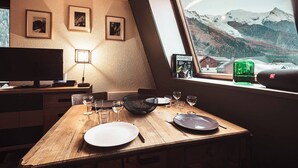 A dining area with a wooden table set for two, a TV on a stand, framed pictures on the wall, and a view of snow-capped mountains through a window. - Vertice - Center Chamonix, Mont-Blanc View (Chamonix-Mont-Blanc)