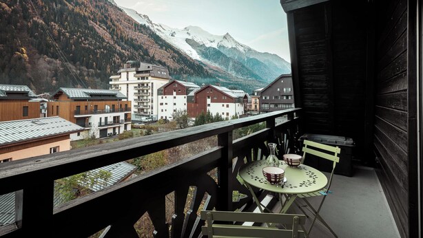 A balcony with a table and chairs, overlooking a mountain village with buildings and snow-capped peaks. - Vertice - Center Chamonix, Mont-Blanc View (Chamonix-Mont-Blanc)