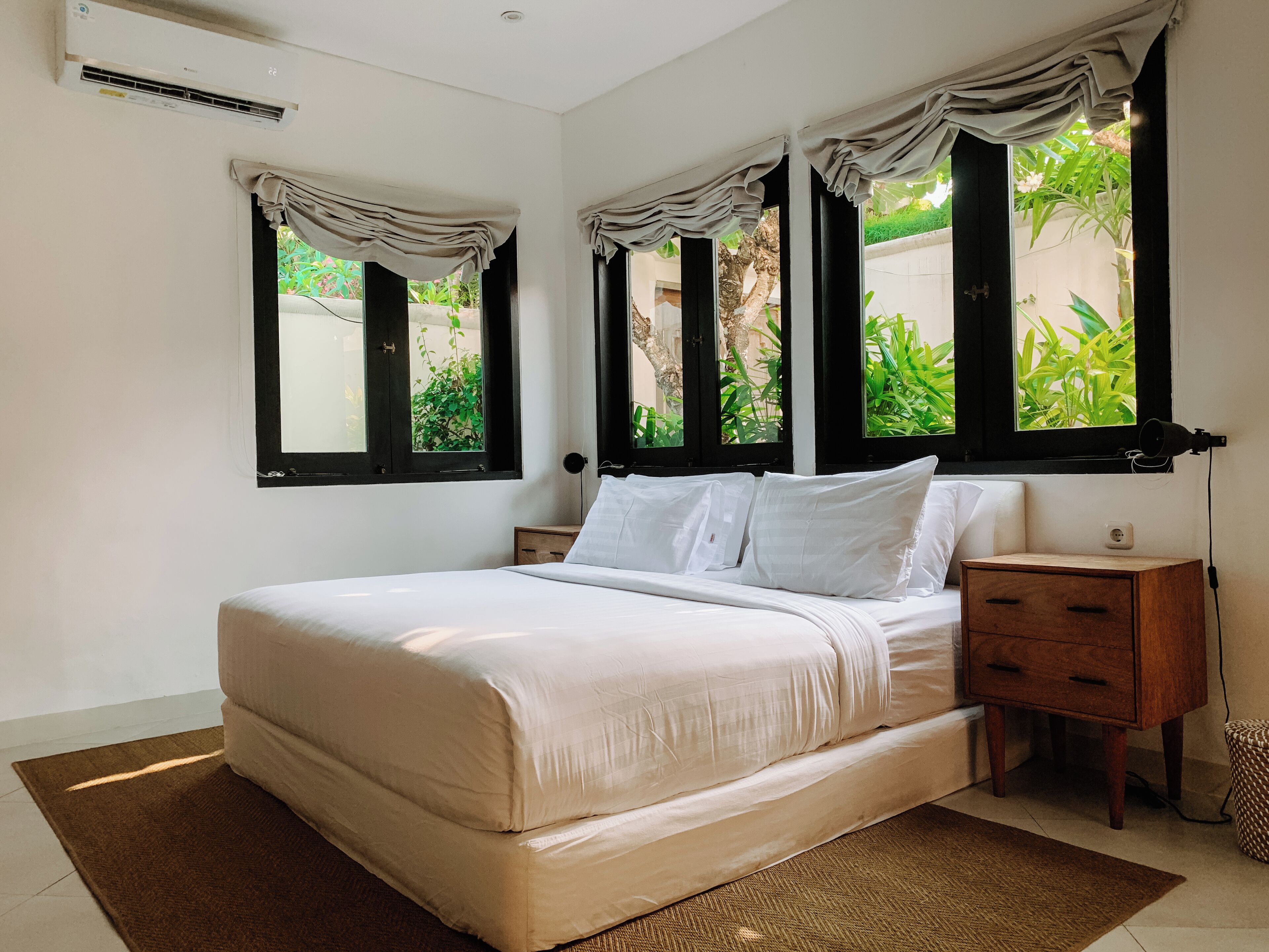 A bedroom with a large bed, two bedside tables, and a nightstand. There are three windows with curtains, and a wall-mounted air conditioning unit.