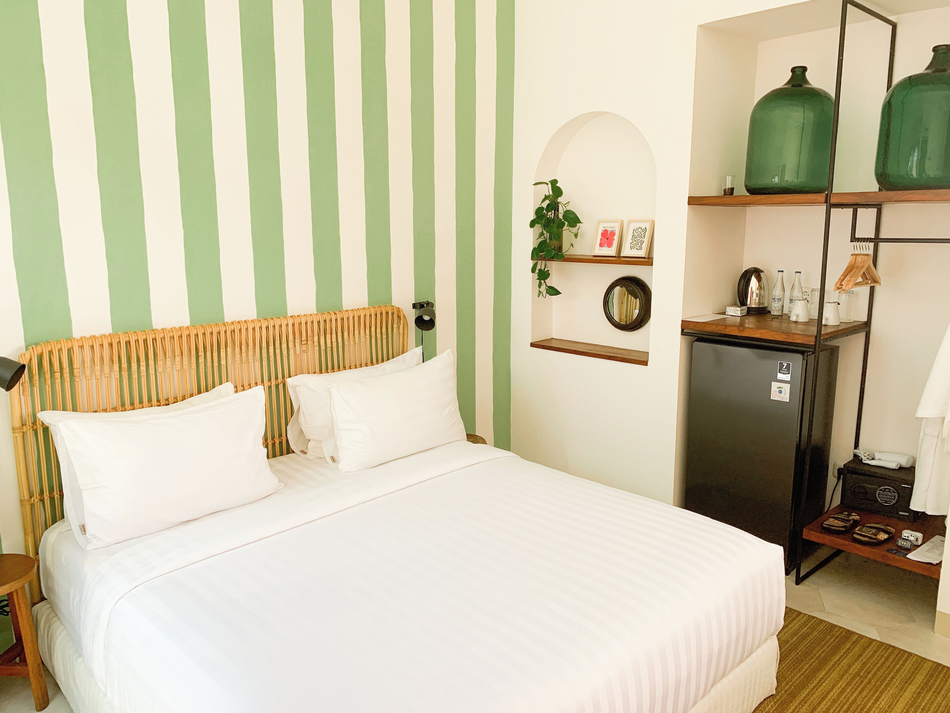 A hotel room with a bed, a small refrigerator, a shelf with decorative items, and a mirror.
