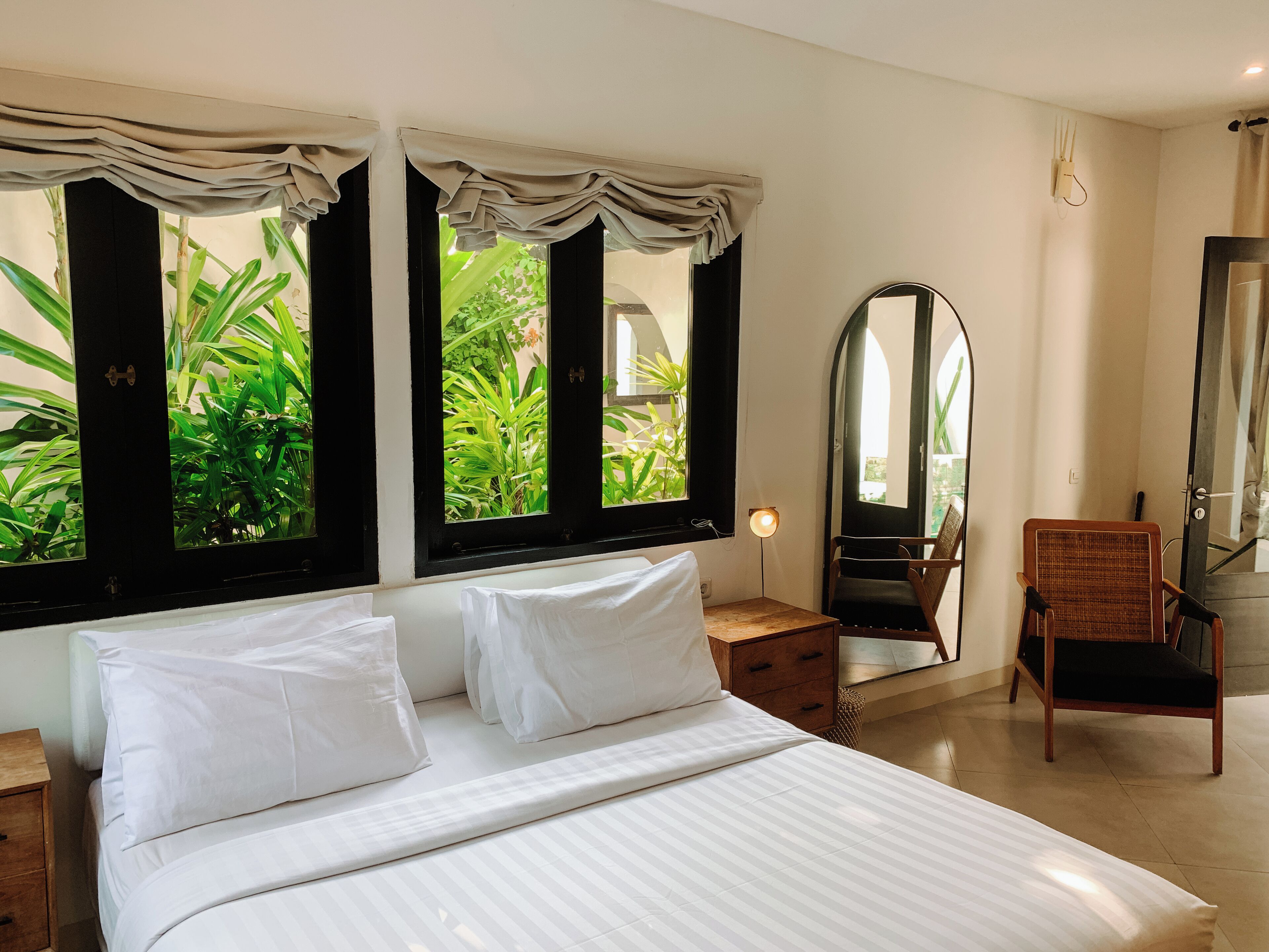 A bedroom with a large bed, two windows with curtains, a bedside table, a chair, a mirror, and a view of greenery outside.