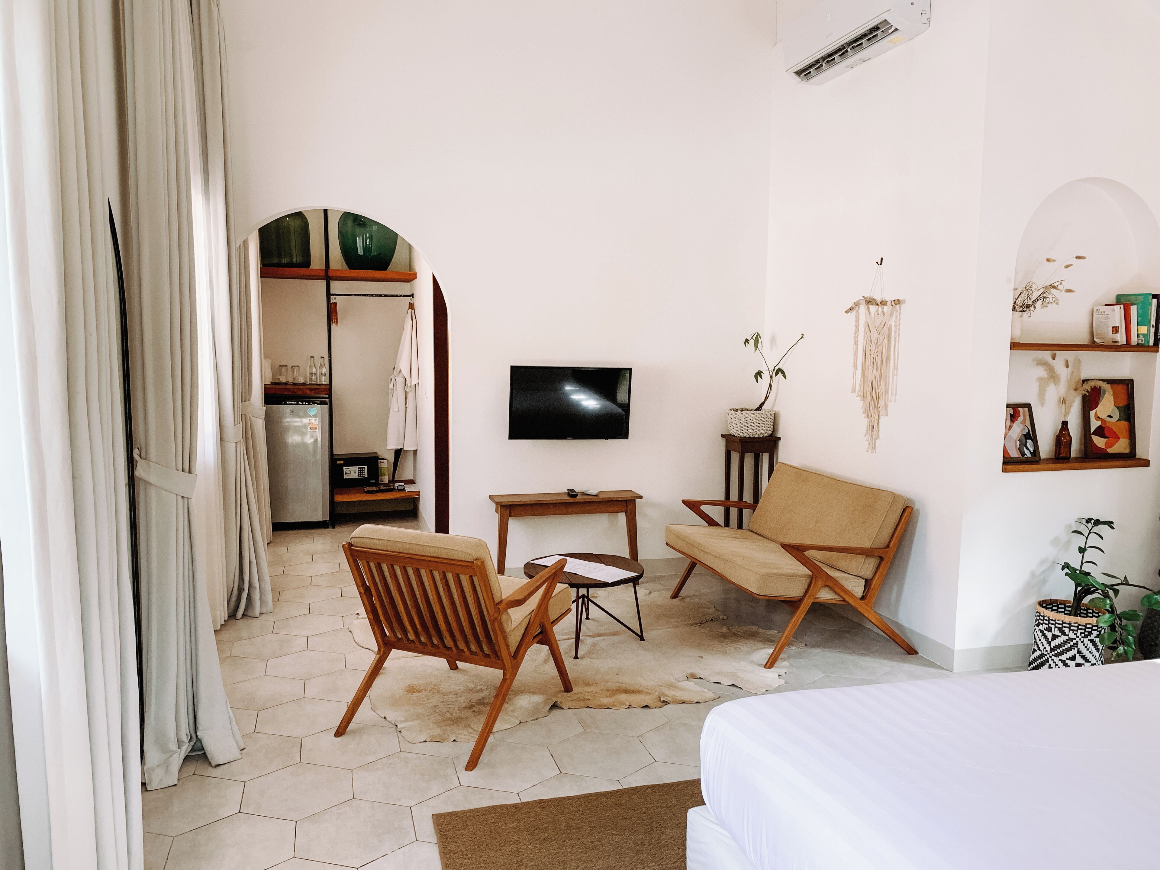 Une chambre d’hôtel moderne comprenant un lit, un canapé, une chaise, une petite table, un téléviseur et un coin cuisine visible à travers une porte ouverte.
