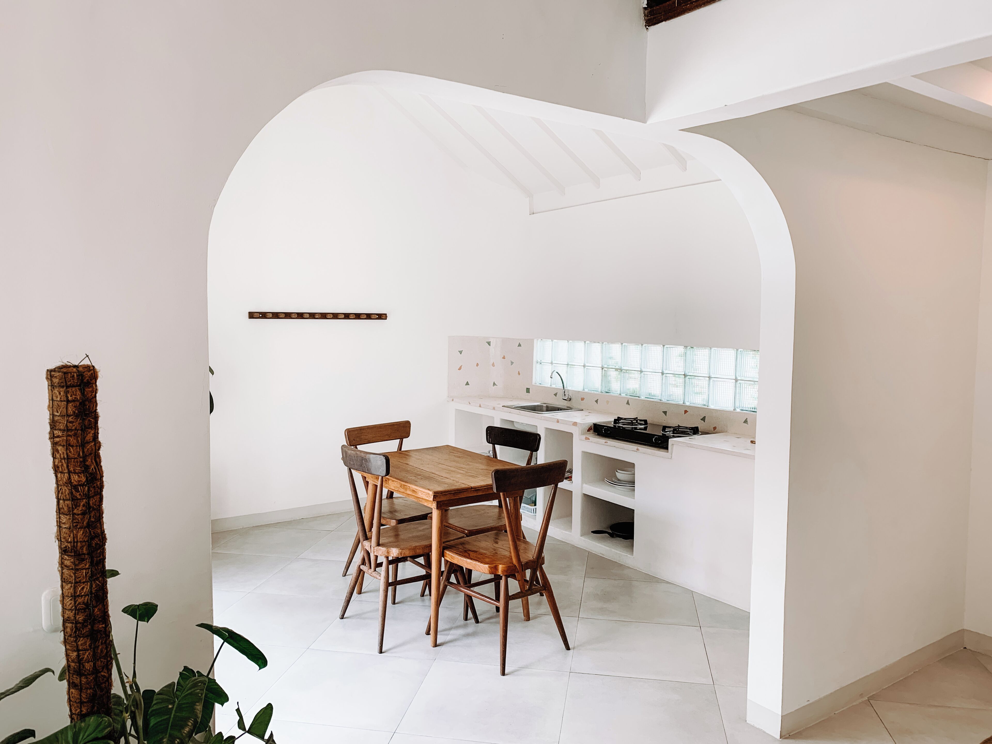 A compact kitchen with a dining area, featuring a white tiled floor, a wooden dining table with four chairs, a white countertop with a stove, and a window with a grid pattern.