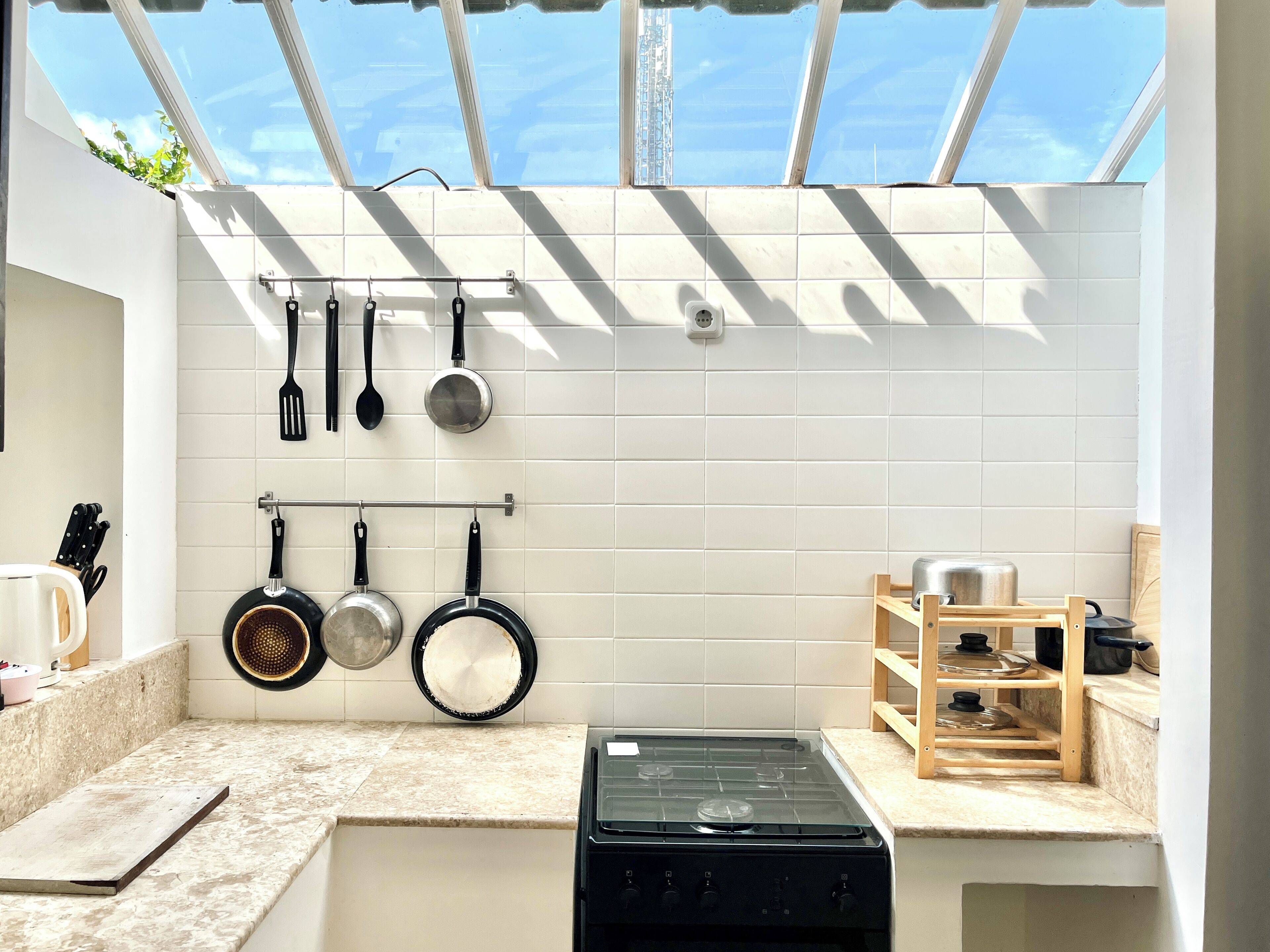 A kitchen with white tiles, a gas stove, hanging pots, and a wooden shelf.