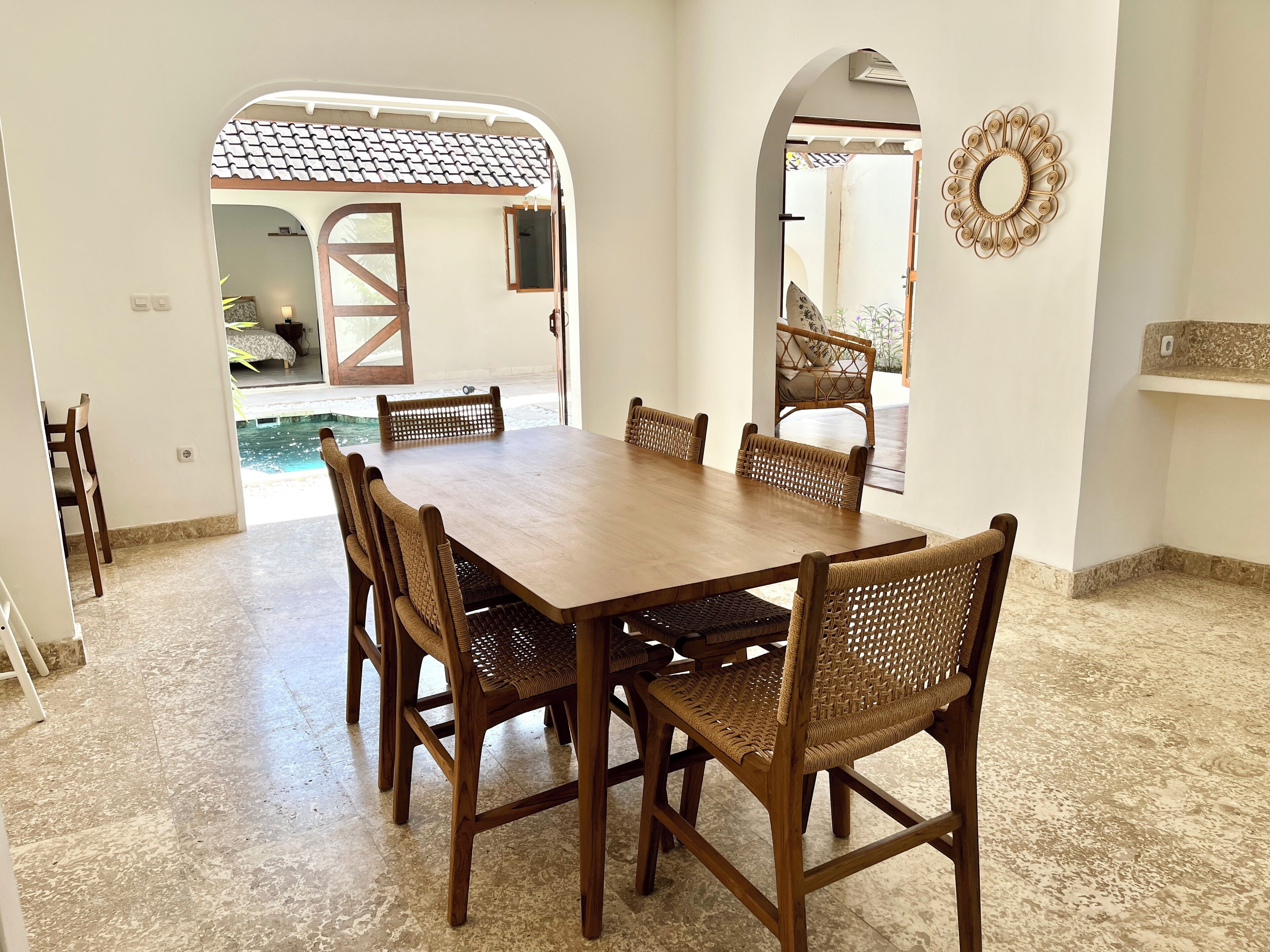 A dining area with a wooden table and woven chairs, a tiled floor, and a view of a garden through an open doorway.