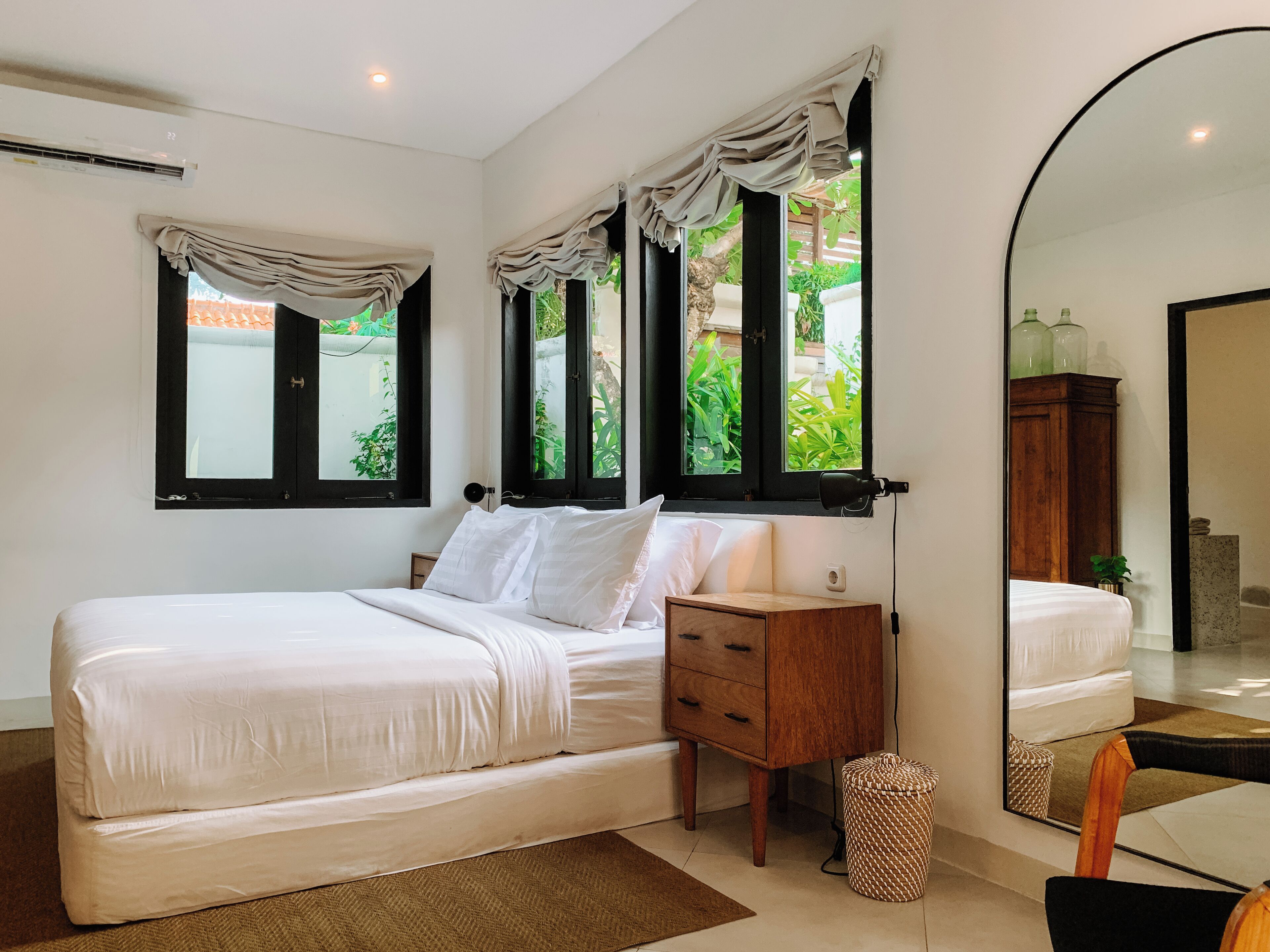 A bedroom with a bed, a nightstand, a mirror, and a window with curtains.