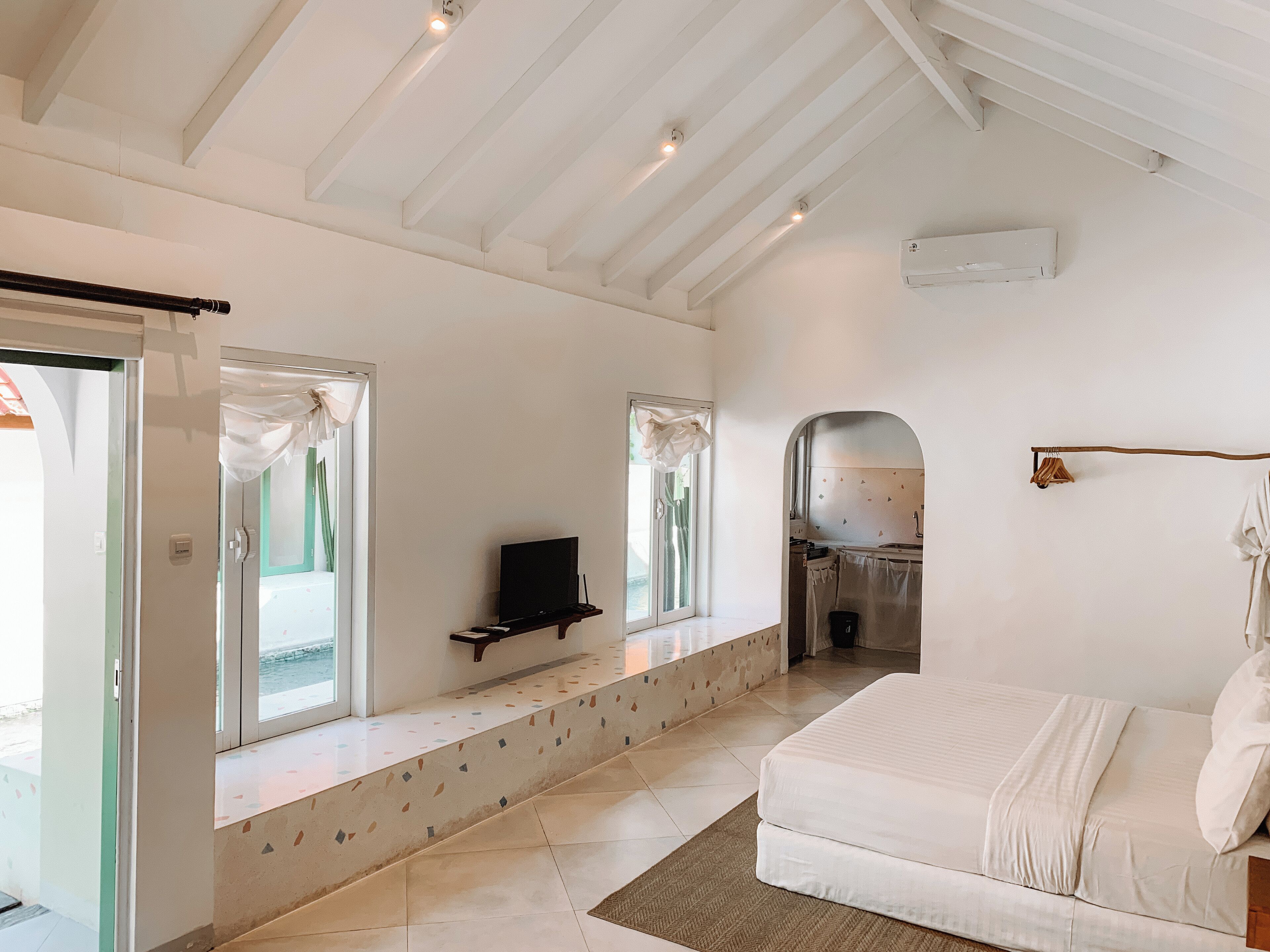 Une chambre à coucher avec un lit, un téléviseur posé sur un meuble, une fenêtre et une salle de bain visible à travers une arche.