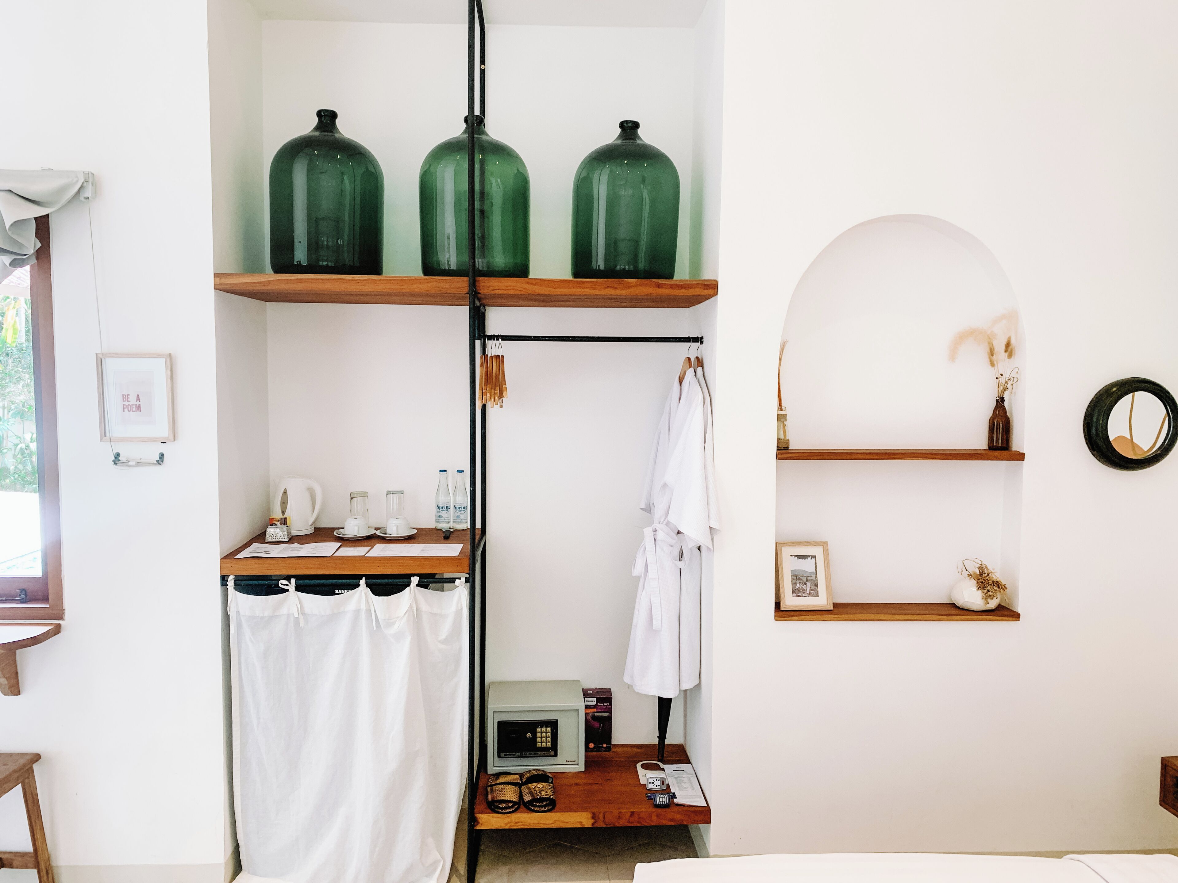 A room with a wooden shelf holding three green glass jars, a white robe hanging on a rack, a small table with a radio, and a mirror on the wall.