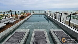 A rooftop infinity pool with a glass edge, surrounded by outdoor seating and a view of the ocean. - Get By Side (João Pessoa)