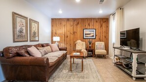 A living room with a brown leather sofa, a wooden accent wall, a television on a white stand, and framed artwork on the wall. - The Erin, 1 BR King Duplex by Velvet Ditch Villas (Oxford)