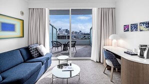 Una habitación de hotel moderna con un sofá azul, una mesita redonda pequeña, un escritorio con una lámpara y vistas de la ciudad a través de puertas correderas grandes.