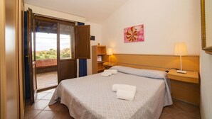 A hotel room with a bed, two bedside lamps, a nightstand, a painting on the wall, and a view through an open door. - Villetta Seaside Sardegna (Villaputzu)