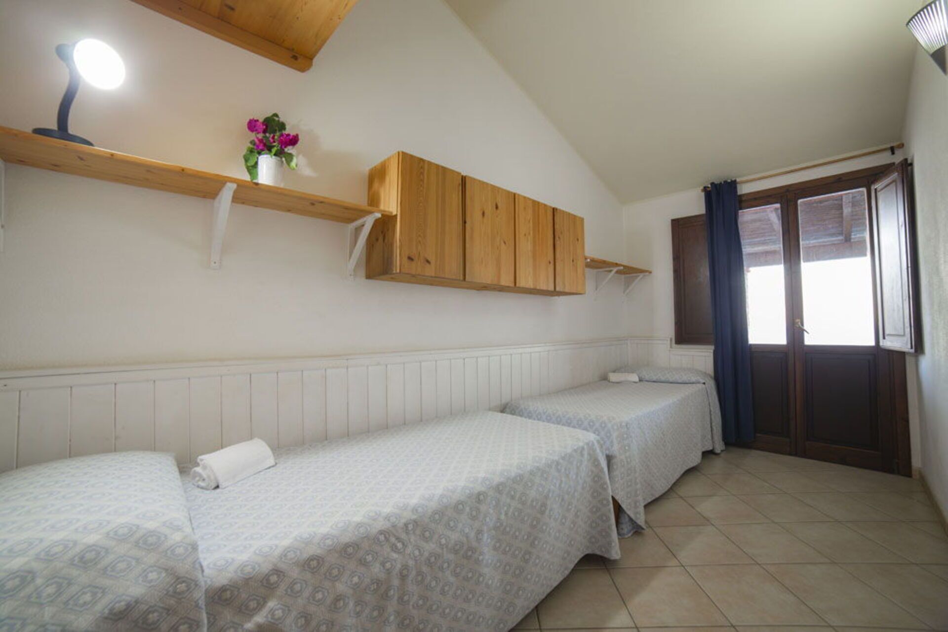 A room with two beds, wooden cabinets, a shelf with flowers, and a window with curtains.