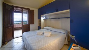 A bedroom with a bed, a desk with a lamp, and a view through a window. - Villetta Seaside Sardegna (Villaputzu)