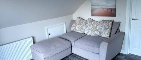 A modern living room with a grey sectional sofa, patterned cushions, a white radiator, and a wooden herringbone floor.