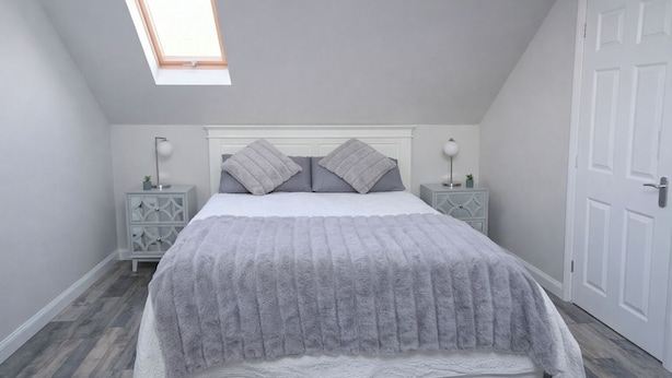 A bedroom with a sloped ceiling, a skylight, a bed with a fluffy gray blanket, two bedside tables with lamps, and a white door. - Lisburn - 1 Bedroom - Sleeps 2 - Street Parking (Lisburn)