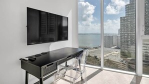 Une chambre moderne dotée d’un grand téléviseur fixé au mur, d’un bureau noir avec une chaise et d’une grande fenêtre donnant sur la ville.