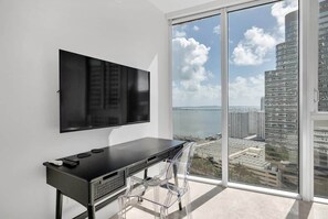 A modern room with a large TV mounted on the wall, a black desk with a chair, and a large window offering a city view. - Icon 2B|2B Fully Remodeled Frontline Bay and Pool (Miami)