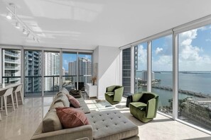 A modern living room with a large window, a sofa, two armchairs, and a dining area with chairs.
